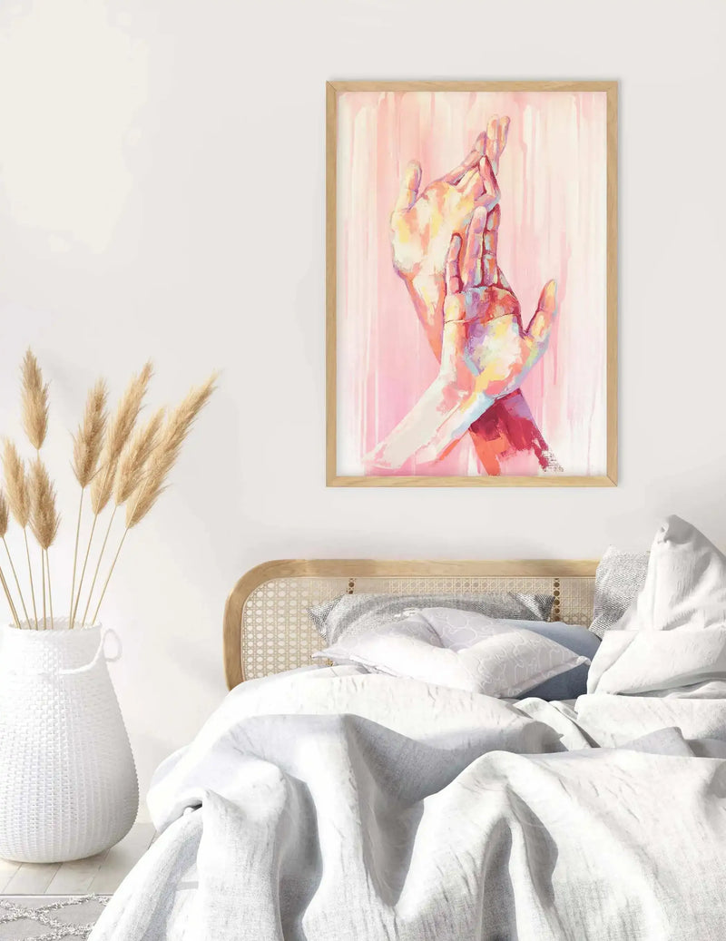"Framed art print displayed above a bed with a wooden cane headboard in a bright bedroom. The print features two hands in an embrace, rendered in an expressive, painterly style with vibrant strokes of pink, red, yellow, and blue against a soft pink background. The room includes a white woven vase with dried pampas grass and white bedding."