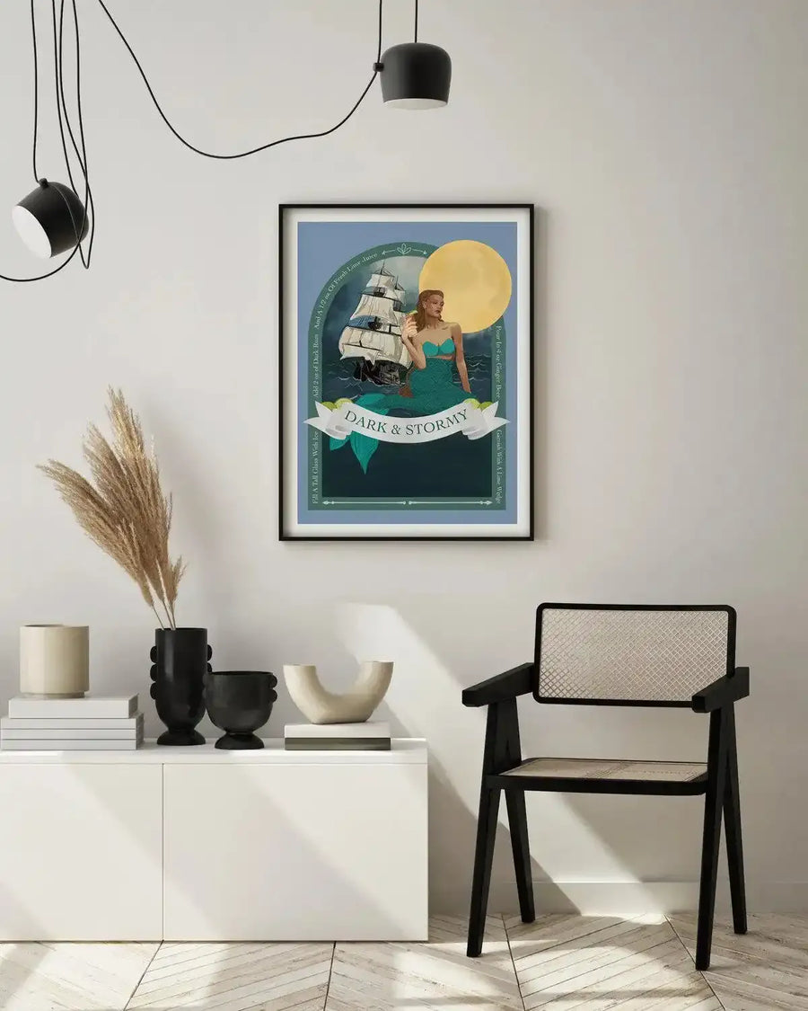 Lifestyle shot of a framed art print displayed above a white console table in a minimalist living room. The print features an illustration of a mermaid with a green tail and teal top, holding a cocktail, next to a sailing ship on a dark blue sea under a large yellow moon. A banner across the bottom reads "DARK & STORMY" with lime slices. The print is in a black frame with a white border. The room includes dried pampas grass in a black vase and a black and cane chair.