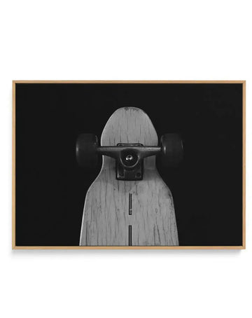 Black and white photograph of a wooden skateboard deck shown front-on against a dark background. The deck features natural wood grain texture with visible vertical striping down the center. A black skateboard truck with metal hardware is mounted horizontally across the upper portion. The image is printed on canvas and displayed in a natural wood frame with a light tan finish.