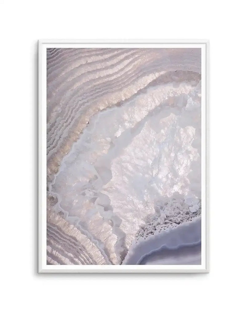 Framed art print titled "Deeper Still" displaying an aerial photograph of concentric curved patterns in soft mauve, lavender, and cream tones. The image shows layered, rippling formations with textured ridges and smooth valleys, suggesting a natural geological or mineral formation. The photograph is mounted in a white frame with a white border, presented against a plain background.