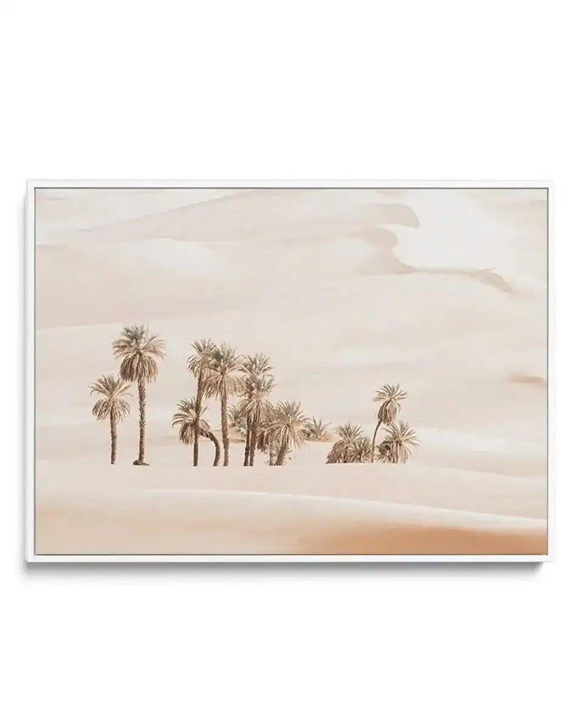 "Framed canvas print of a desert oasis with a white frame. The artwork features a cluster of palm trees in varying heights, with sandy-coloured trunks and fronds, set against a backdrop of rolling sand dunes in soft beige and pale peach tones under a bright, diffused sky."