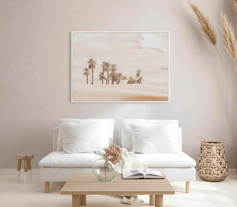 Lifestyle shot of a minimalist living room with a framed canvas art print displayed above a white sofa. The print features a desert landscape with numerous palm trees scattered across rolling sand dunes under a soft, muted sky, rendered in warm, sandy tones. The room includes a light wood coffee table with a glass vase holding dried flowers, a woven basket, and a small wooden stool.