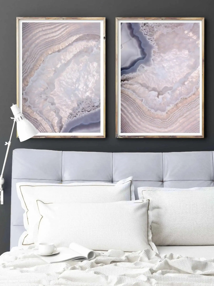 Lifestyle shot of a bedroom featuring two framed art prints displayed above a grey upholstered headboard. The prints feature abstract close-up photography of agate stone formations with swirling patterns in pale lavender, white, and soft beige, displayed in natural wood frames with white borders. The bed is dressed with white pillows and a white textured throw, with a white desk lamp on the left.