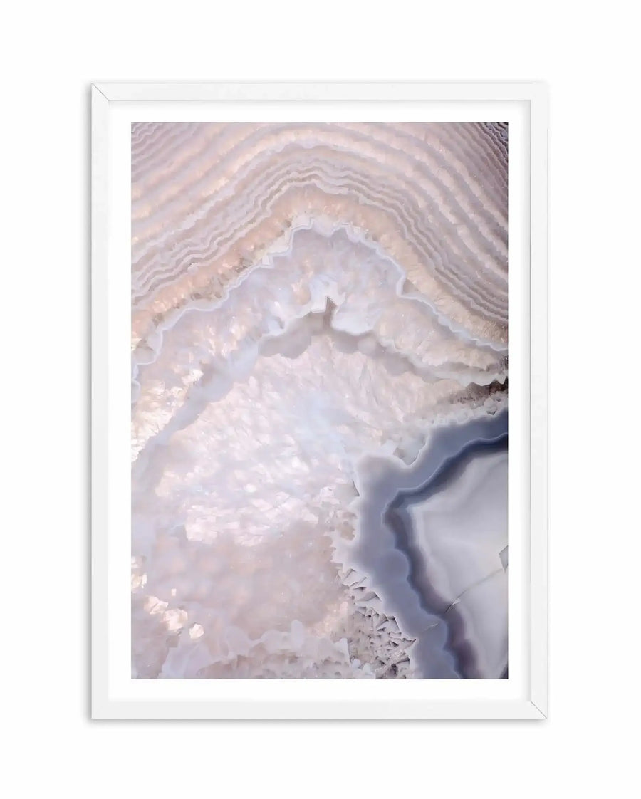 Framed art print featuring an aerial photograph of concentric agate layers in soft mauve, lavender, and cream tones. The naturally banded mineral formation shows delicate curved striations with a central white cavity, creating organic geode-like patterns. White wooden frame with white border surrounding the vertical composition.