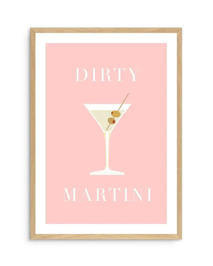 Art print featuring a minimalist illustration of a dirty martini cocktail on a pale pink background, displayed in a natural oak frame with white border. The martini glass is filled with a pale yellow liquid and garnished with two green olives and one red olive on a pick. The words "DIRTY" and "MARTINI" are written in a light cream serif font above and below the illustration.