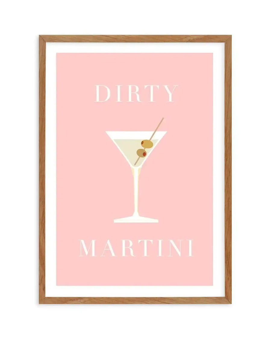 Art print featuring a minimalist illustration of a dirty martini cocktail on a soft pink background, displayed in a natural oak frame with white border. The martini glass is white with a pale yellow liquid and two green olives and one red pimento on a toothpick. The words "DIRTY" and "MARTINI" are written in a light cream serif font above and below the illustration.