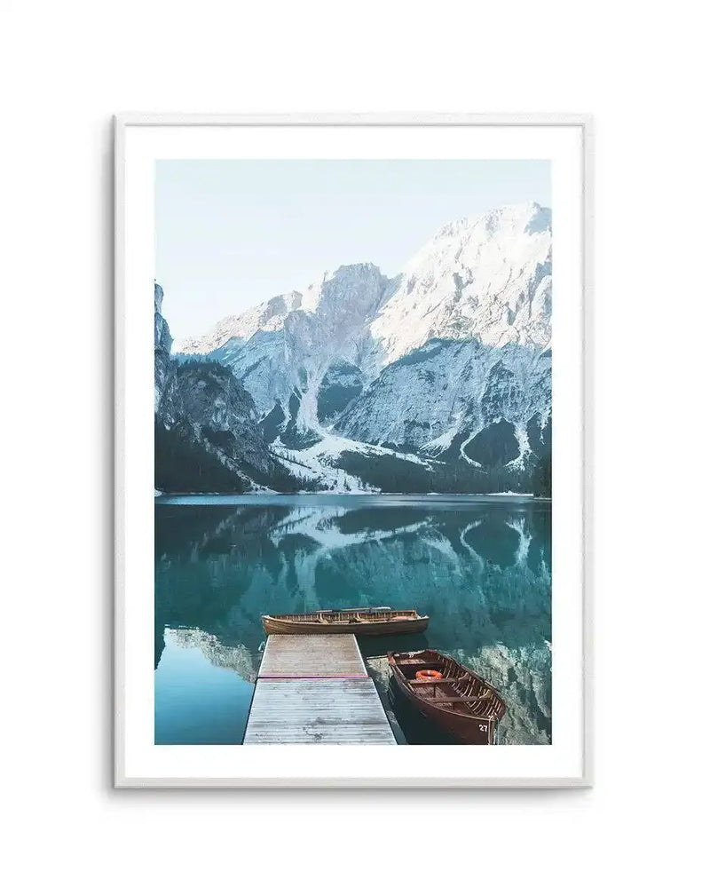 Framed photograph of a serene mountain lake scene with a white frame and white border. The composition features snow-capped mountains reflecting in the still, turquoise water, with a wooden pier extending into the lake where two wooden boats are moored. The sky is a clear, pale blue.