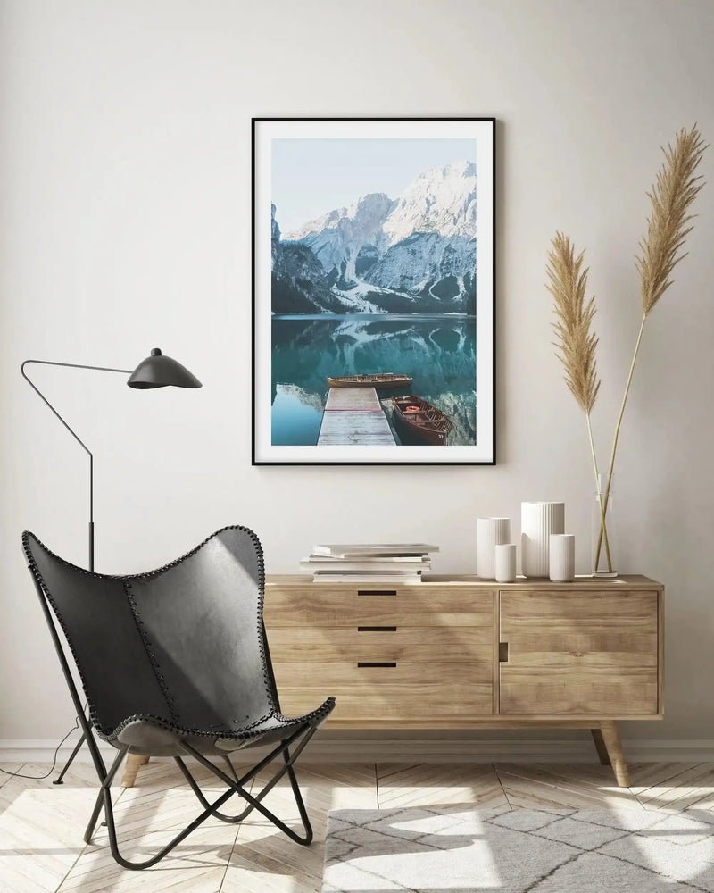 Large framed art print displayed above a wooden sideboard in a minimalist living room. The print features a photograph of a serene mountain lake with snow-capped peaks reflecting in the turquoise water, a wooden pier, and two wooden boats. The print has a black frame and white border. The room includes a black leather butterfly chair, a black floor lamp, a stack of books, white ceramic vases, and dried pampas grass.