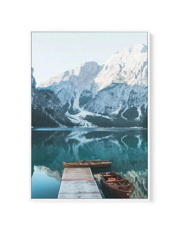 Framed canvas art print featuring a serene alpine lake landscape. The photograph shows snow-capped Dolomite mountains with steep rocky peaks reflected in turquoise-blue water. In the foreground, a wooden dock extends into the lake with two rustic wooden rowboats moored alongside. Dense evergreen forests line the lake shores beneath the mountains. The canvas is mounted in a light gray frame with white border. Cool blue and white tones dominate the composition, creating a peaceful, winter mountain atmosphere.