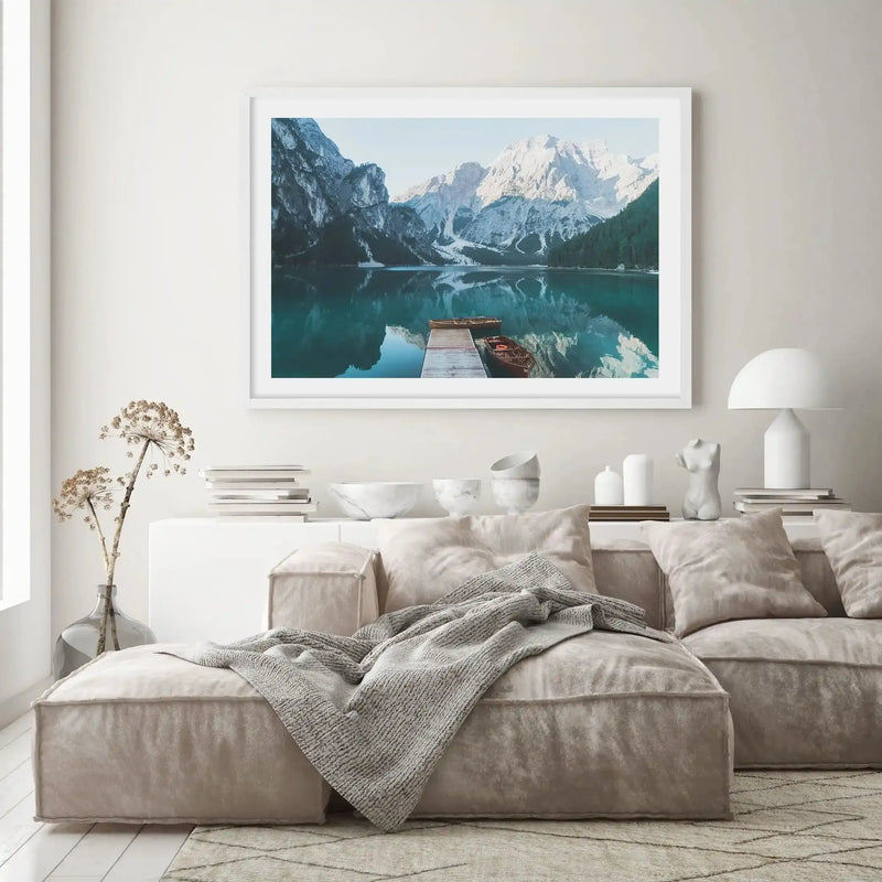 Large framed art print displayed above a white console table in a minimalist living room. The print features a photograph of a serene mountain lake with snow-capped peaks reflecting in the turquoise water, with a wooden pier and two boats in the foreground, with a white frame and white border. The room includes a large cream sectional sofa with a grey knitted throw, a white table lamp, and dried floral arrangements.