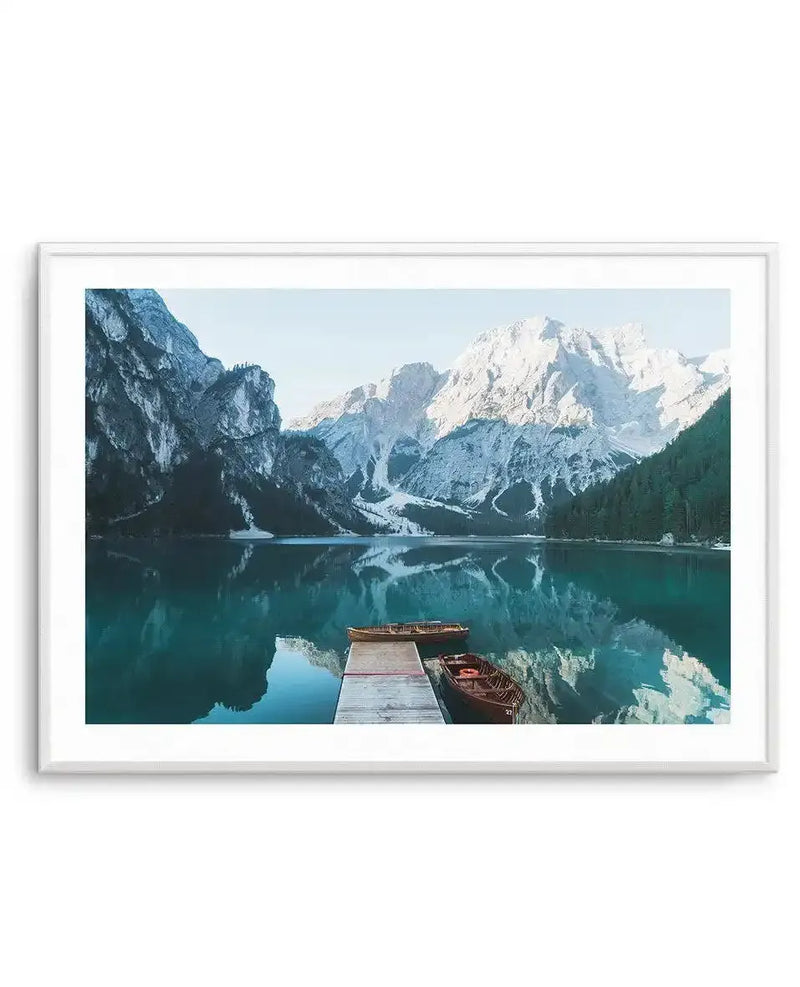 Framed art print displaying a serene Alpine lake landscape. A wooden dock with two red rowboats extends into turquoise water that mirrors towering snow-capped mountains and dramatic grey limestone cliffs. Dense evergreen forests line the lake shores beneath a pale blue sky. The photograph captures cool tones of teal, white, and slate grey. Presented in a white frame with white border.