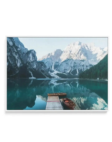 Framed canvas print featuring a serene photographic landscape of the Dolomites mountain range reflected in a clear, turquoise lake. The composition includes a wooden dock extending into the water with two traditional wooden boats moored alongside, set against a backdrop of towering, snow-capped peaks under a bright sky. The print is displayed in a white frame.