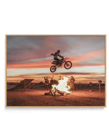 Framed canvas art print titled "Doom At Dusk" by Tim Harris featuring a motocross rider airborne on a dirt bike against a dramatic sunset sky in shades of orange, pink, and grey. Below, spectators stand around a blazing bonfire in a desert landscape at dusk. The scene captures a stunt performance with industrial bleachers visible on the left side. Natural wood frame with canvas print mounted with white border.