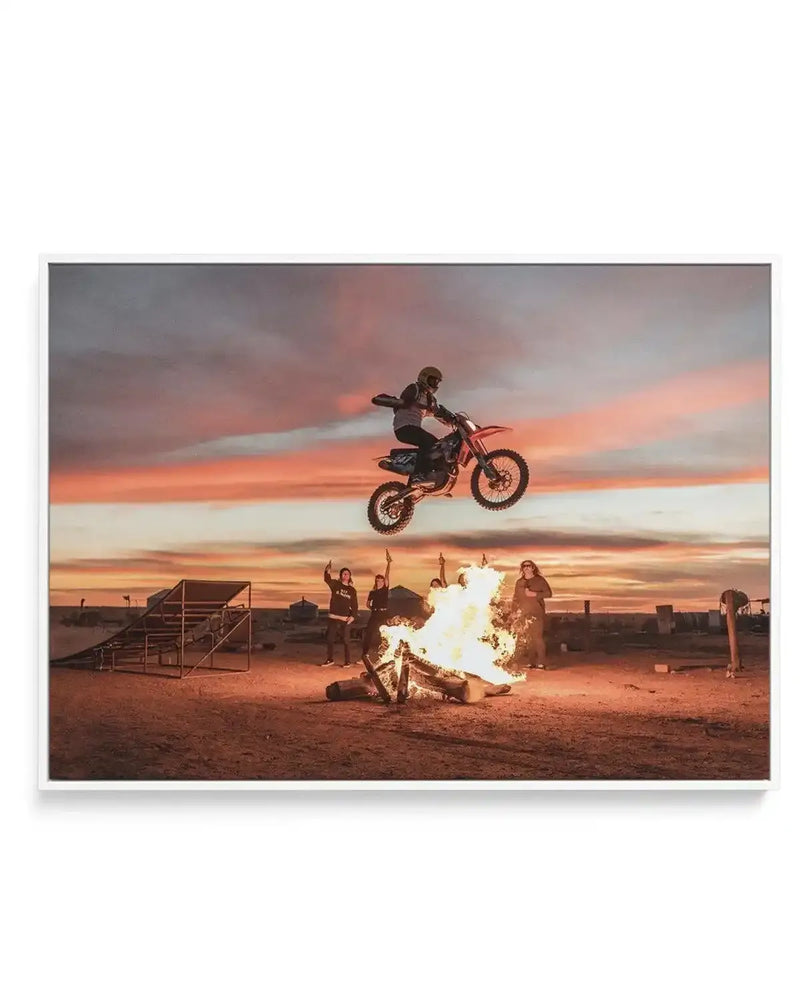 Framed canvas art print featuring a dramatic photograph titled "Doom At Dusk" by Tim Harris. The image captures a motocross rider performing an airborne stunt on a dirt bike against a striking sunset sky filled with orange, pink, and purple hues. Below, spectators stand near a blazing bonfire on desert terrain. A metal ramp structure is visible on the left. The photograph has warm, dramatic lighting with the sun setting behind the scene. The canvas is mounted in a gray frame with a white border.