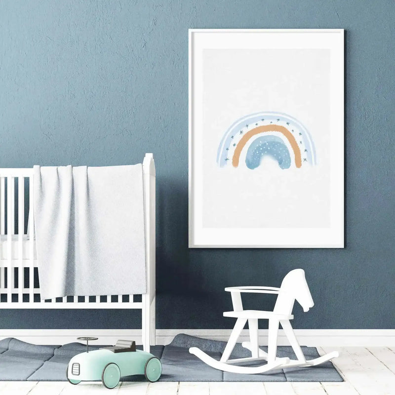 Lifestyle shot of a baby's nursery with a framed art print displayed above a white crib. The print features a dreamy watercolour rainbow in muted blue and soft orange with small blue star accents on a white background, displayed in a white frame with a white border. The room has textured blue walls, a light wood floor, a grey rug, a white rocking horse, and a light blue toy car.