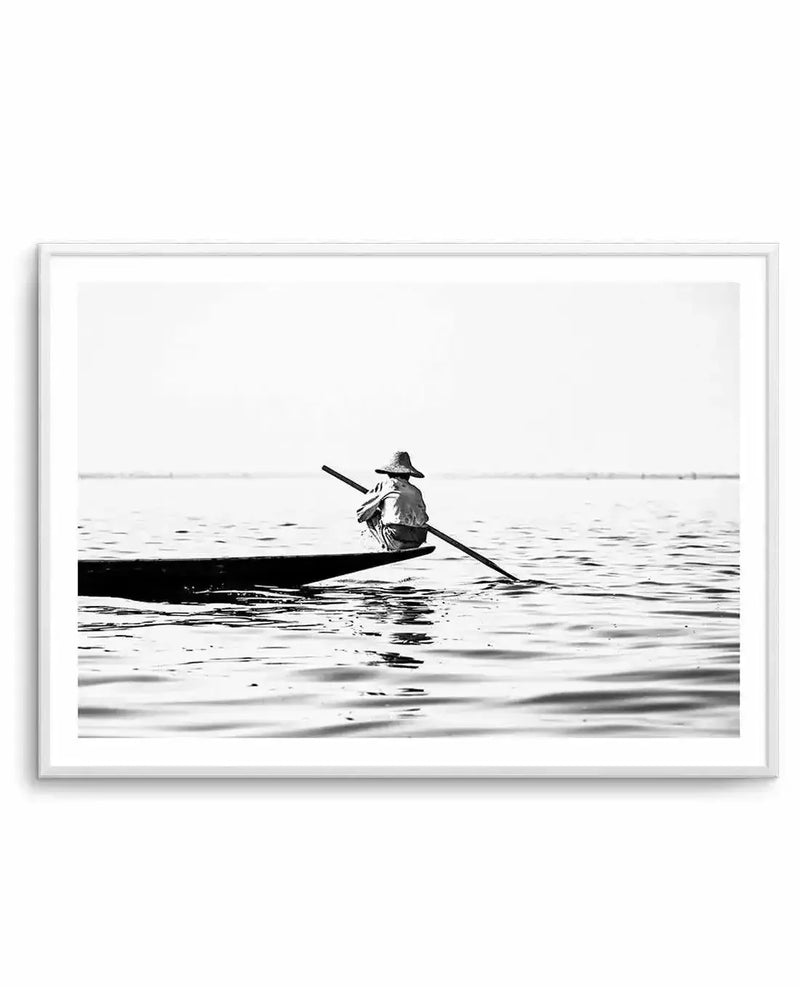 Framed black and white photograph of a person in a conical hat rowing a small, narrow boat on a calm body of water, with a white frame and white border. The person is seen from behind, positioned in the middle of the boat, holding a long oar that extends into the water. The water shows subtle ripples, and a faint horizon line separates it from a bright, featureless sky.