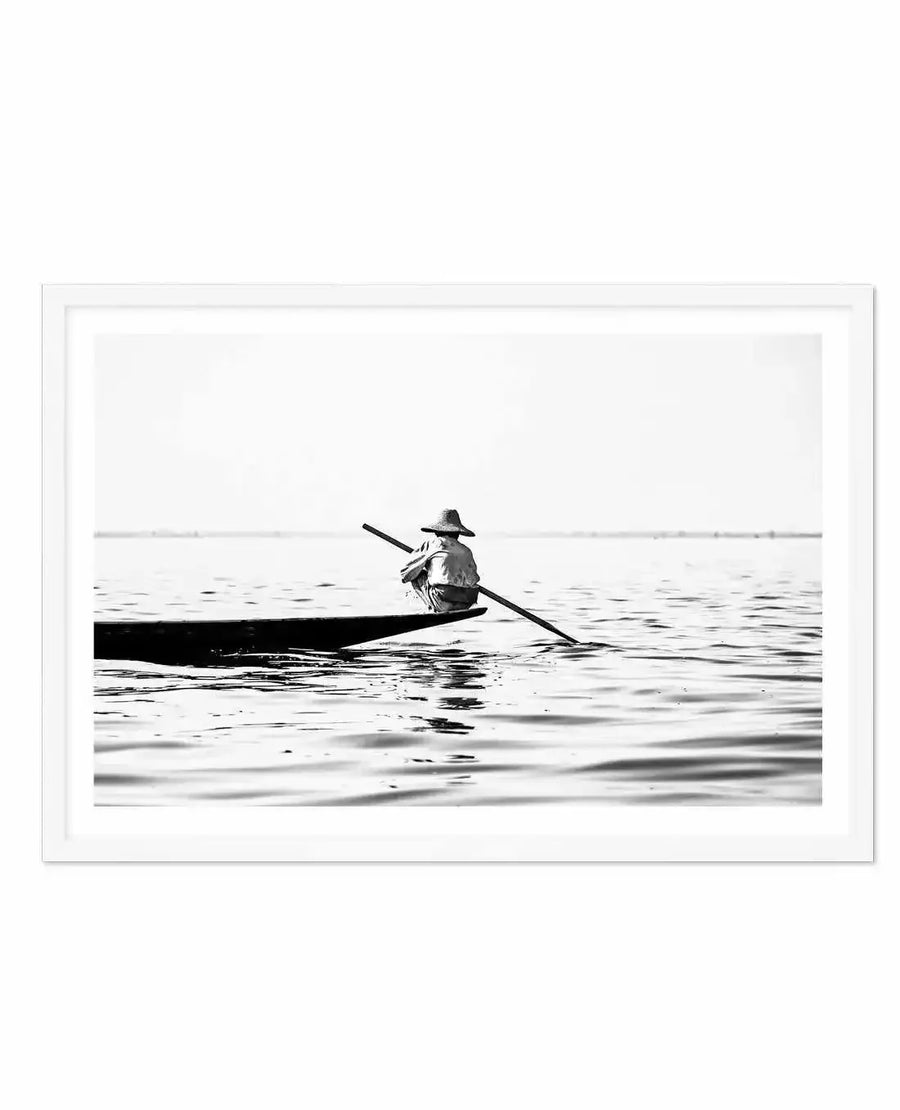 Framed black and white photograph of a lone person in a boat on calm water, displayed in a white frame with white border. The person, wearing a conical hat, is seen from behind, sitting in the boat and holding a long oar. The composition features a wide expanse of water with subtle ripples, and a distant horizon line under a bright sky, creating a serene and minimalist mood.