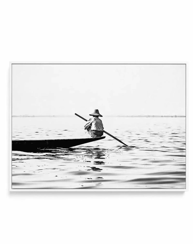 Black and white framed canvas art print featuring a solitary figure in a conical hat paddling a narrow boat on still water. The boatman leans forward with a long oar, captured in a minimalist composition with calm, rippled water in the foreground and a distant, hazy horizon line. The photograph has a serene, contemplative mood with high contrast between the dark boat and figure against the lighter water and sky. The canvas is mounted in a thin gray frame with a white border.