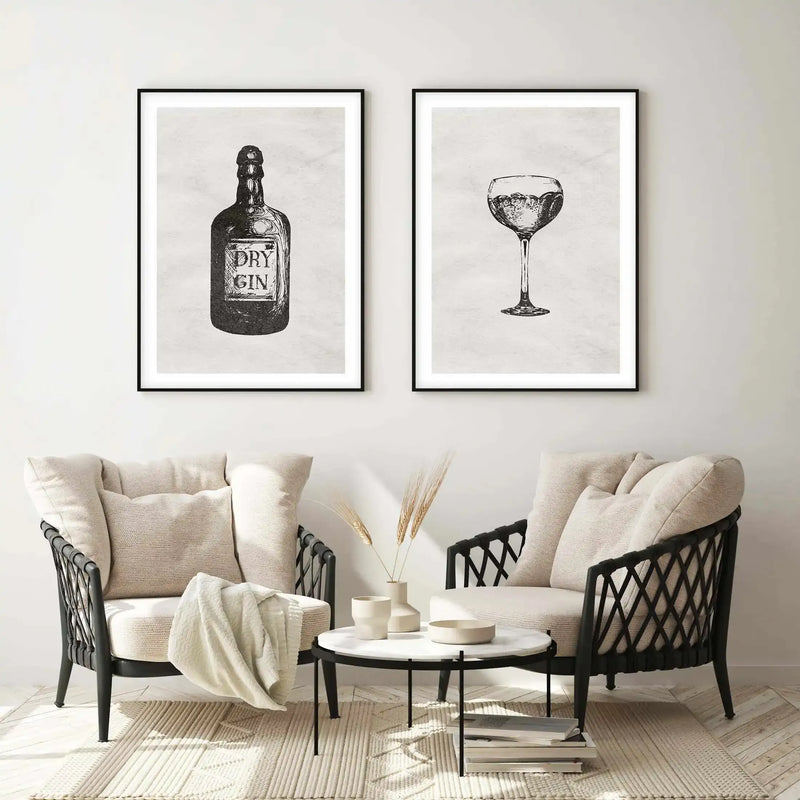 Lifestyle shot of two framed art prints displayed above two armchairs in a minimalist living room. The left print features a black and white illustration of a "Dry Gin" bottle, and the right print features a black and white illustration of a gin glass with liquid and bubbles. Both prints have a black frame and white border. The room includes two cream upholstered armchairs with black woven frames, a round marble coffee table with black legs, a cream textured rug, and dried wheat stalks in ceramic vases.