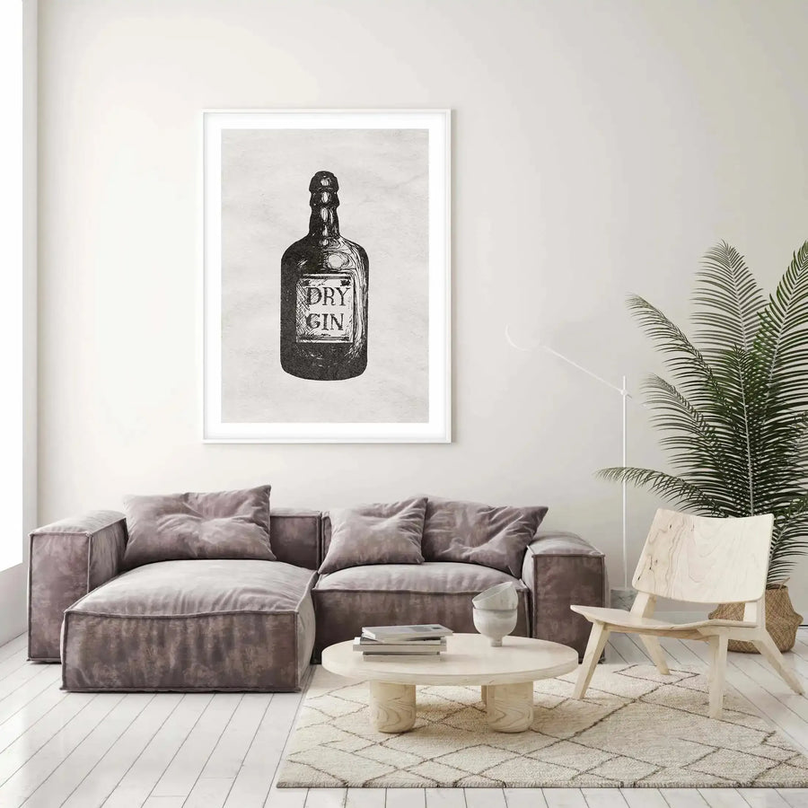 Lifestyle shot of a minimalist living room featuring a large framed art print of a "Dry Gin" bottle above a grey sectional sofa. The print is a black and white illustration of a vintage-style gin bottle with the words "DRY GIN" on the label, displayed in a white frame with a white border. The room includes a light wood coffee table with books and a ceramic bowl, a light wood chair, a tall floor lamp, and a large green palm plant in a woven basket.