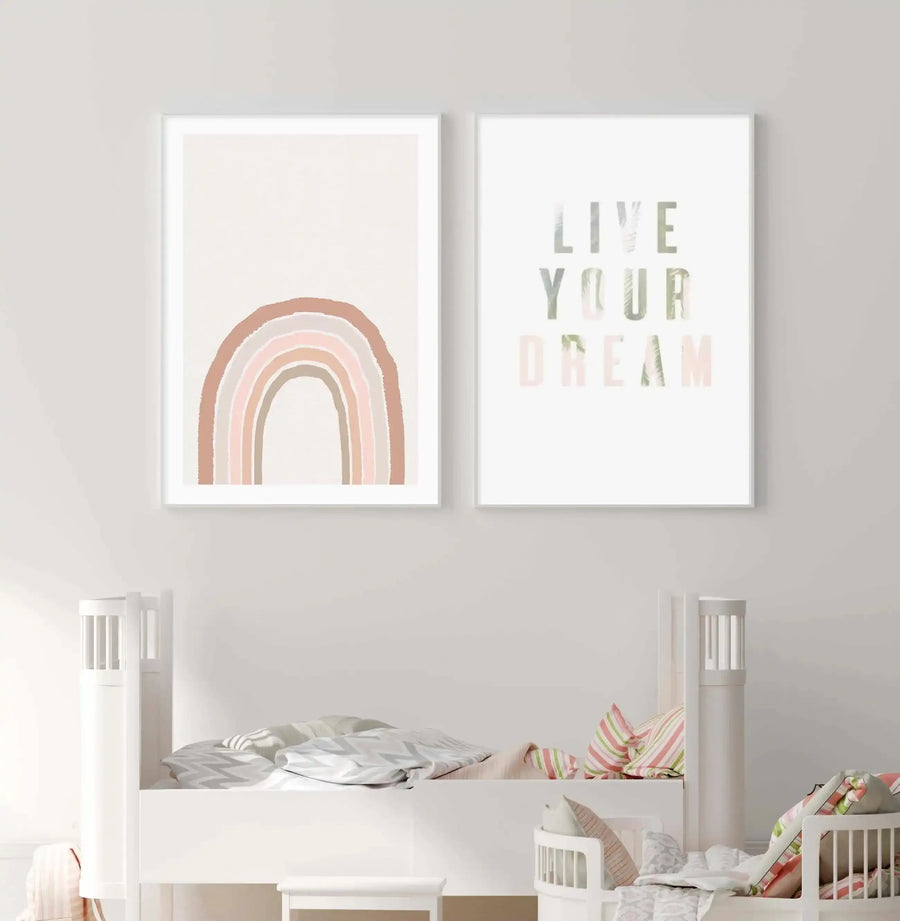 Lifestyle shot of a child's bedroom featuring two framed art prints above a white bed. The left print depicts a hand-painted rainbow in muted tones of dusty rose, terracotta, and beige on a cream background, with a white frame and white border. The right print features the words "LIVE YOUR DREAM" in a distressed, light green font with a subtle leaf pattern, also in a white frame with a white border. The bed is adorned with patterned bedding in grey, white, pink, and green stripes.