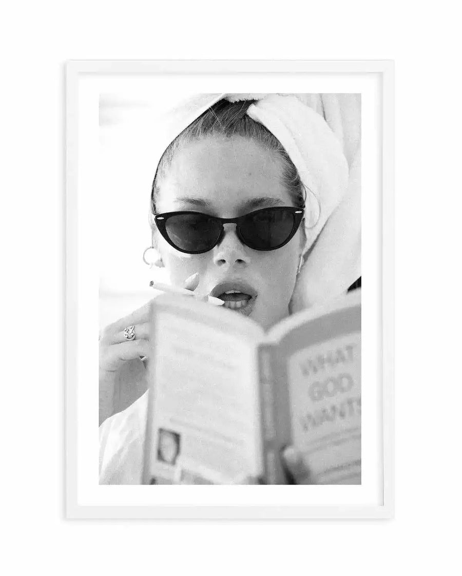 Framed black and white photograph of a woman with a towel wrapped around her head, wearing cat-eye sunglasses, smoking a cigarette, and reading a book titled "WHAT GOD WANTS". The composition captures her from the chest up, with a white frame and white border.