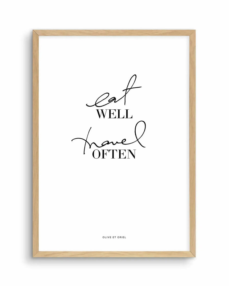 Framed art print with natural wood frame displaying black script and serif typography on white background. Text reads "eat well" in flowing cursive script, "WELL" in serif capitals below, followed by "travel" in script and "OFTEN" in capitals. Small text at bottom reads "OLIVE ET ORIEL". Minimalist design with balanced composition centered on white paper with white border within frame.