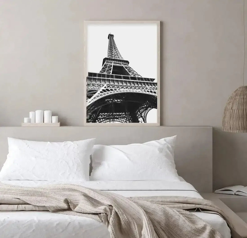 Black and white art print of the Eiffel Tower photographed from below, showing the iconic iron lattice structure rising dramatically against a white sky. The composition captures the tower's geometric framework and intricate metal details from an upward angle. The print is displayed in a light wood frame with white border, hung above a bed with white pillows and a cream knit throw blanket in a minimalist bedroom setting. A woven pendant lamp and white candle holder are visible on the bedside surface.