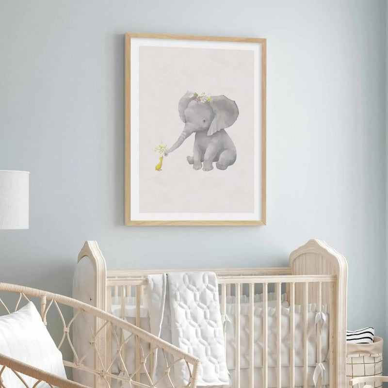 Nursery:
Framed art print displayed above a light wood crib in a nursery with light blue walls. The print features a watercolour illustration of a baby elephant with a crown of flowers on its head, offering a small bouquet of yellow flowers to a little yellow duckling. The print is displayed in a natural wood frame with a white border. The crib has white bedding and a quilted white blanket, and a white lamp is visible on the left.