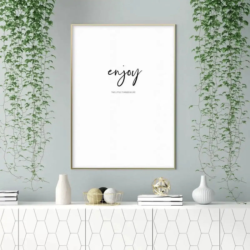 Large framed art print displayed above a white console table in a minimalist living room. The print features the word "enjoy" in black script font with "THE LITTLE THINGS IN LIFE" in smaller black sans-serif text below, set against a clean white background with a thin gold frame. The room includes two cascading green vine plants, a white ceramic vase, a gold wire basket with decorative balls, and a white ribbed vase.