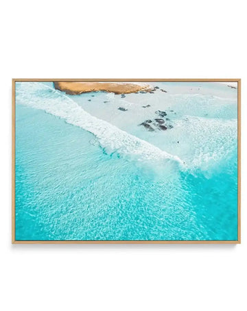 Gallery-wrapped canvas print featuring an aerial photograph of a vibrant turquoise ocean with white waves breaking onto a light sand beach. The clear water reveals dark rocks scattered near the shore, and a small figure appears to be surfing or swimming in the waves. The print is displayed in a natural wood frame.