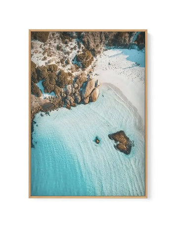 Aerial photograph framed in natural wood showing a pristine turquoise beach cove surrounded by rugged tan and brown rocky cliffs with white snow patches. The shallow aquamarine water meets a curved white sand beach, with dark rock formations scattered in the shallow waters. The composition captures the dramatic coastal landscape from directly above, displaying the striking contrast between the jewel-toned water, pale sand, and weathered cliff face. Framed in light natural wood with white border.