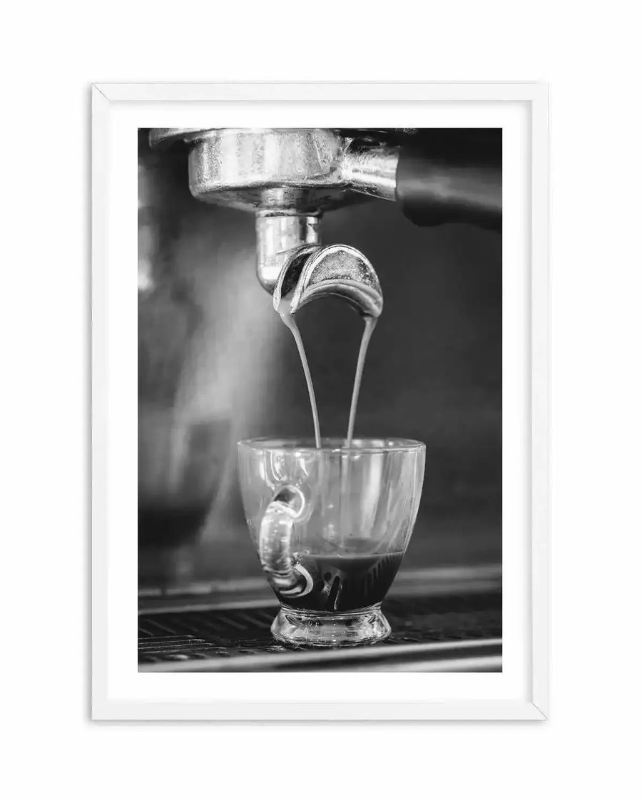 Framed black and white photograph of espresso being extracted from a coffee machine into a clear glass cup, with a white frame and white border. The composition captures the moment two streams of espresso pour from the portafilter into the cup, creating a rich dark liquid with a light crema on top.