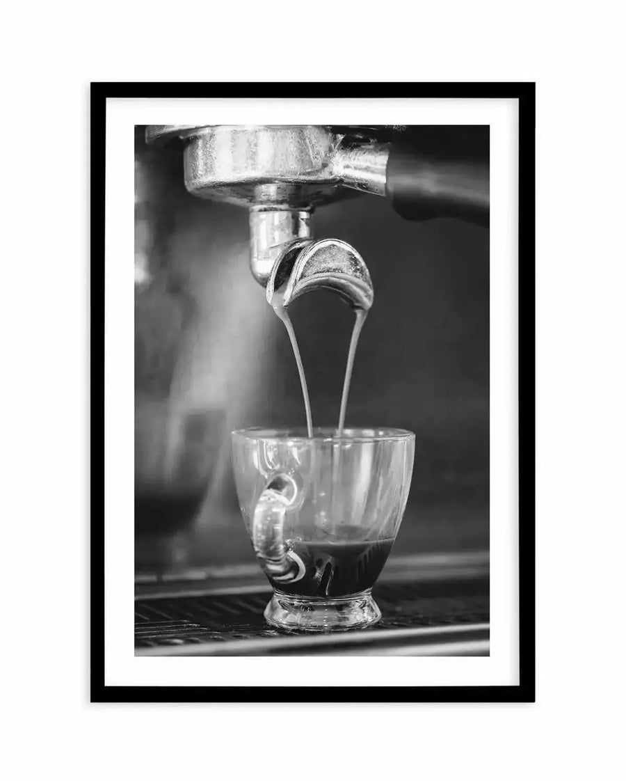 Framed black and white photograph of espresso being extracted from a portafilter into a clear glass cup, with a black frame and white border. The composition captures the moment two streams of dark espresso with light crema pour into the cup, creating a dynamic and close-up view of the coffee-making process.