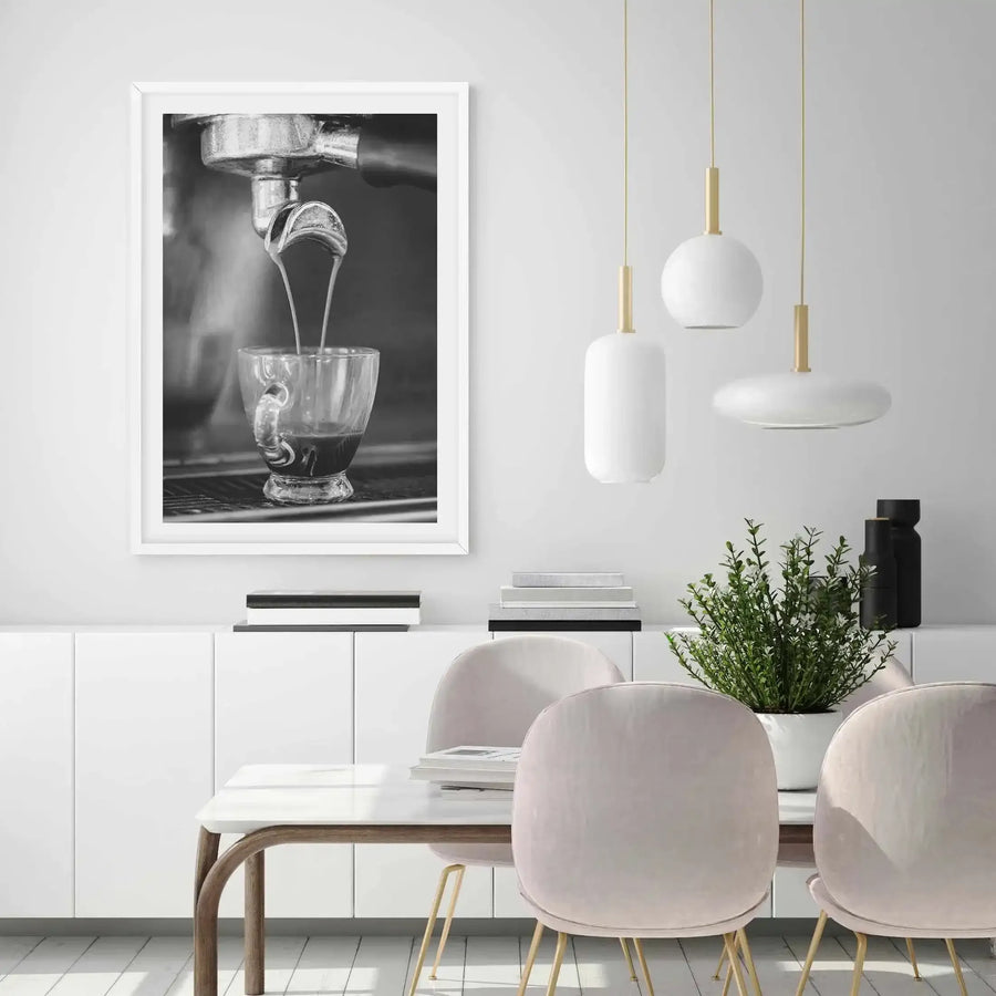 Lifestyle shot of a minimalist dining room featuring a framed black and white photograph of an espresso machine pouring coffee into a clear glass mug. The print is displayed on a white wall above a long white cabinet. The room includes a white dining table with light pink velvet chairs, three modern white pendant lights with gold accents, and a potted green plant on the cabinet.