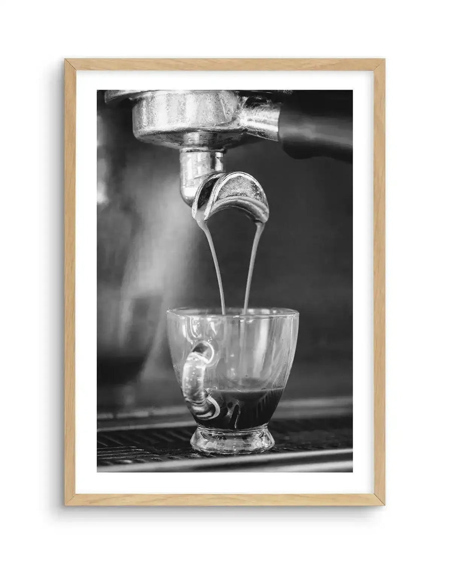 Framed black and white photograph of espresso being brewed into a clear glass cup, with natural wood frame and white border. The composition captures two streams of espresso pouring from a portafilter into the cup, with steam rising in the background.