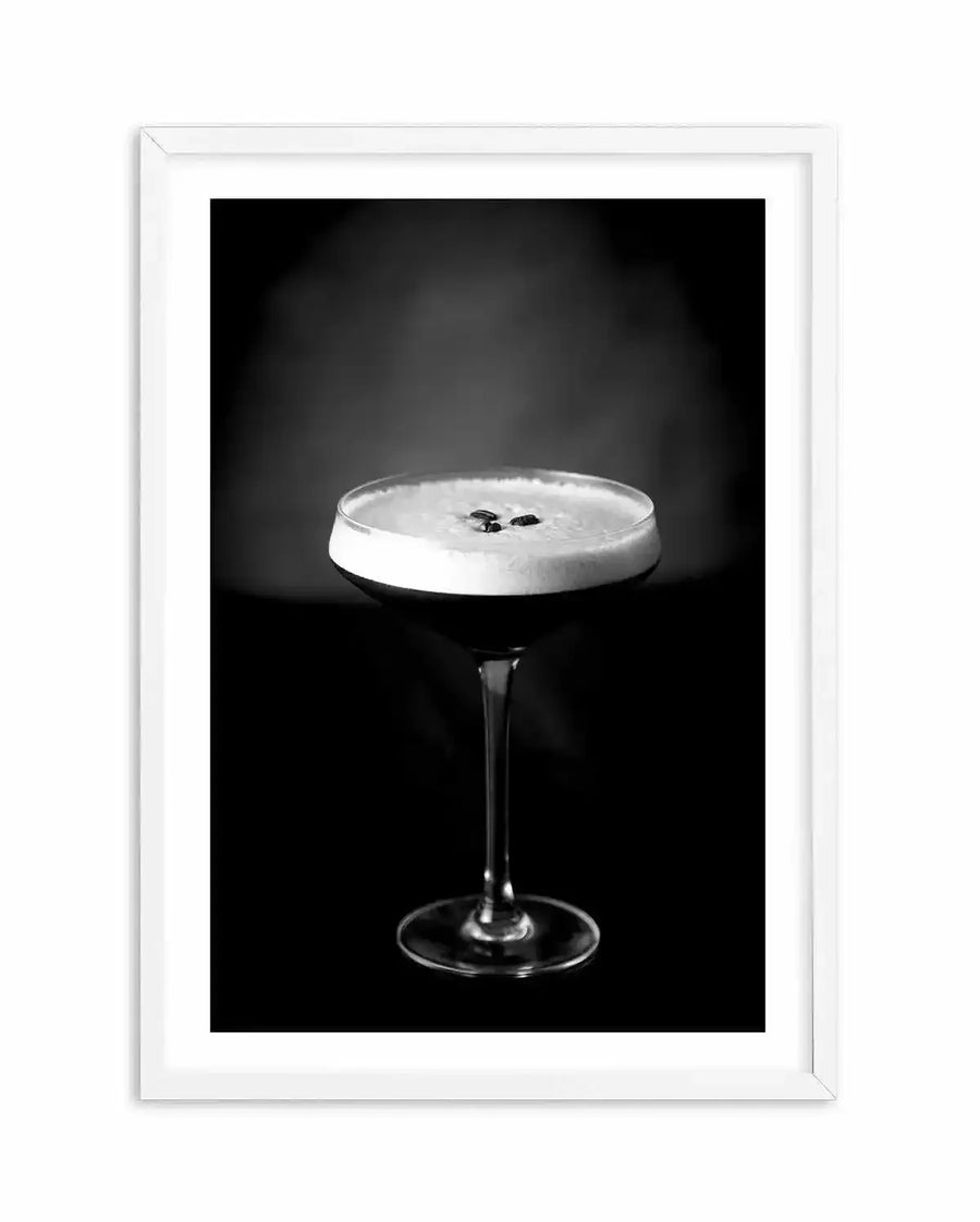 Black and white photograph of an Espresso Martini cocktail in a coupe glass against a dark background. The drink features a distinctive creamy white foam layer topped with two dark coffee beans. The glass has a thin stem and sits centered in the composition. The photograph is mounted in a white wooden frame with a white border, creating a gallery-style presentation. The high-contrast monochromatic aesthetic emphasizes the texture of the foam and the clarity of the glassware.