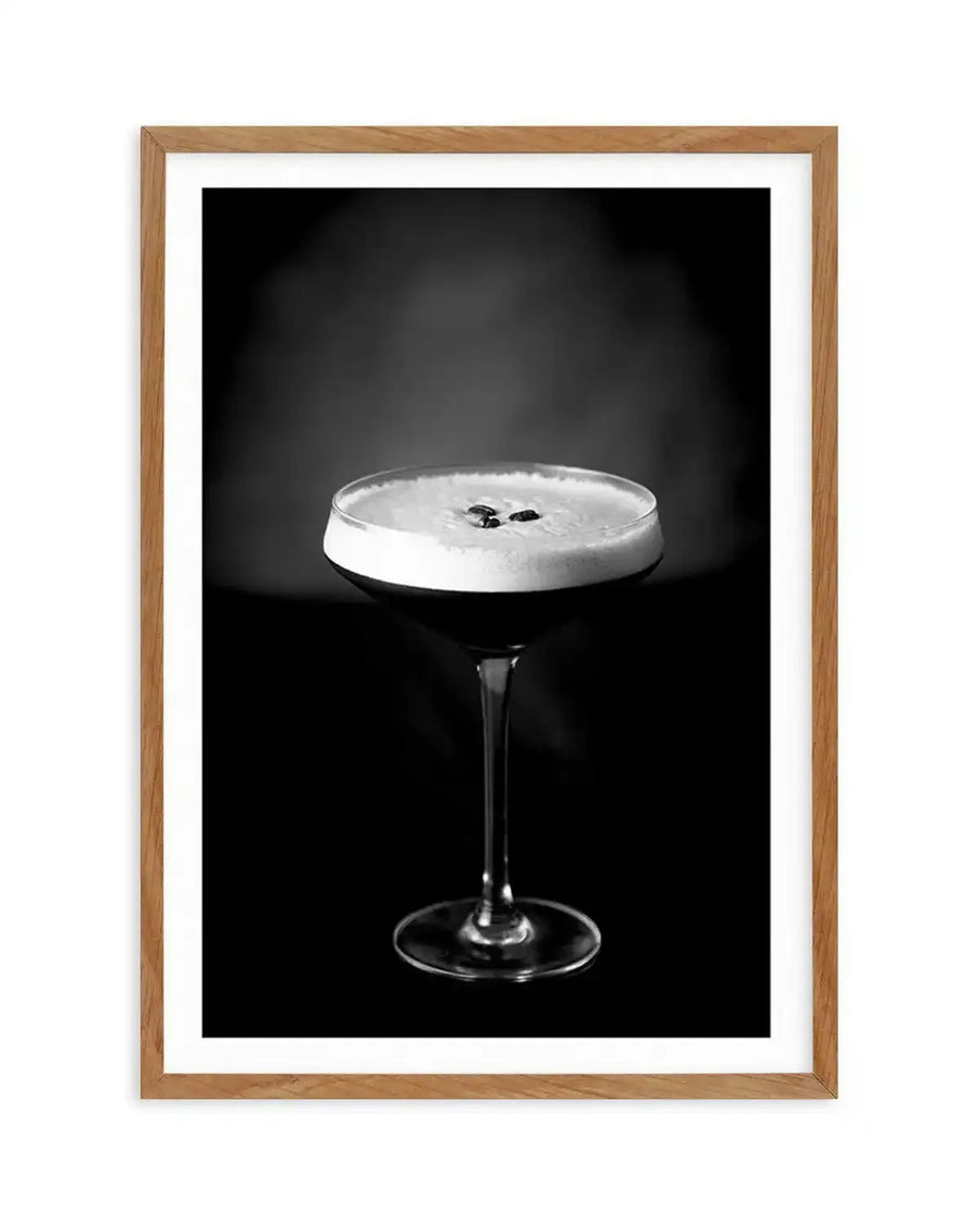 Black and white photograph of an Espresso Martini cocktail in a coupe glass against a dark background. The drink features a creamy white foam top garnished with two dark coffee beans. The glass has a thin stem and round base. The image is mounted in a natural wood frame with a white border.