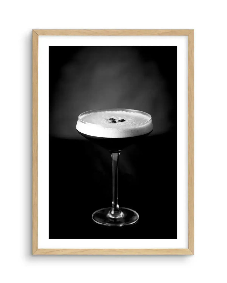 Black and white photograph of an Espresso Martini cocktail in a coupe glass with a thick foam head and two coffee beans on top, set against a dark background. The image is displayed in a natural wood frame with a white border.