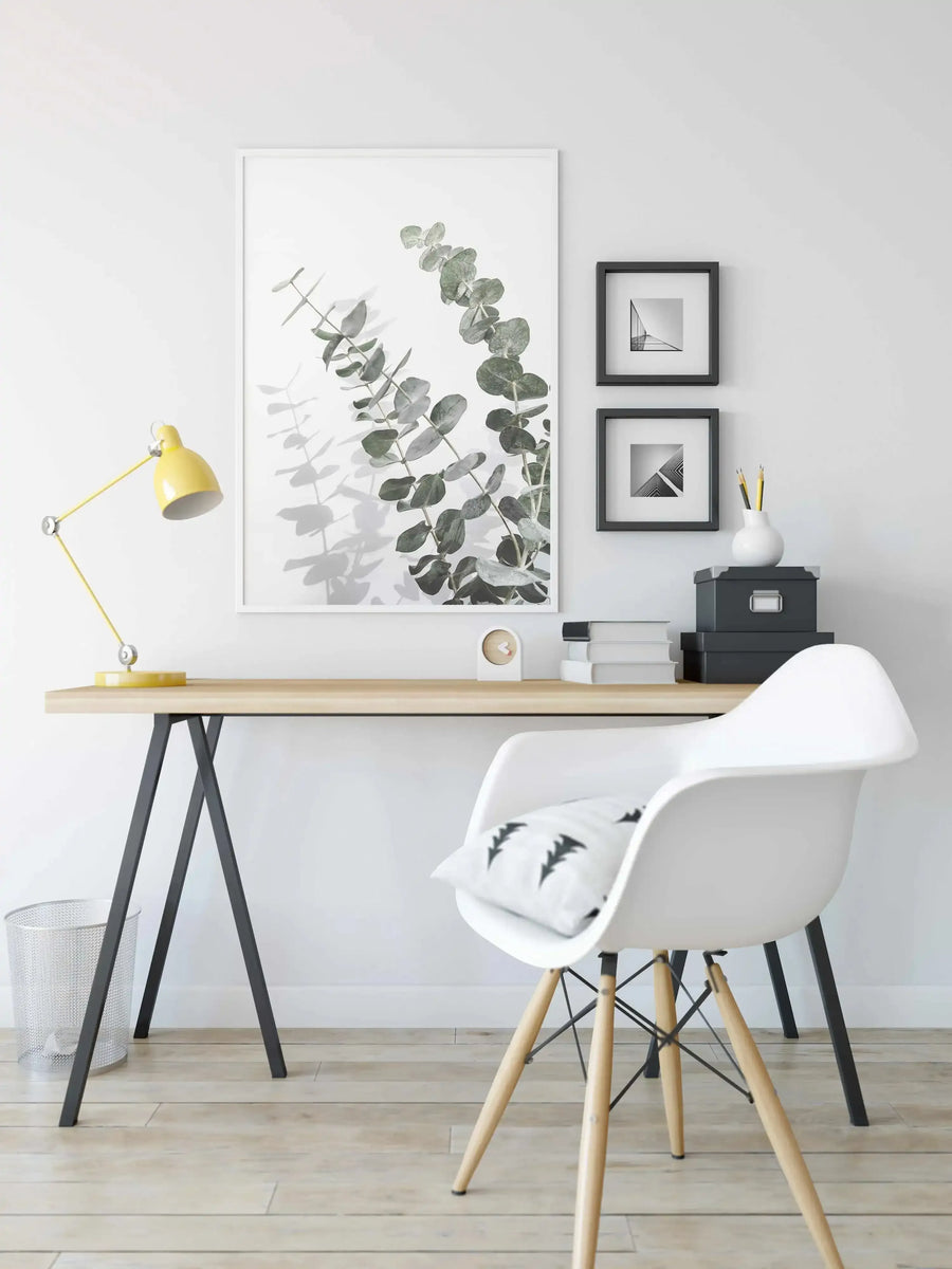 Framed art print featuring a botanical watercolor illustration of eucalyptus branches with rounded green leaves in muted sage and gray tones on white background, displayed in a light wood frame with white border above a minimalist desk workspace.