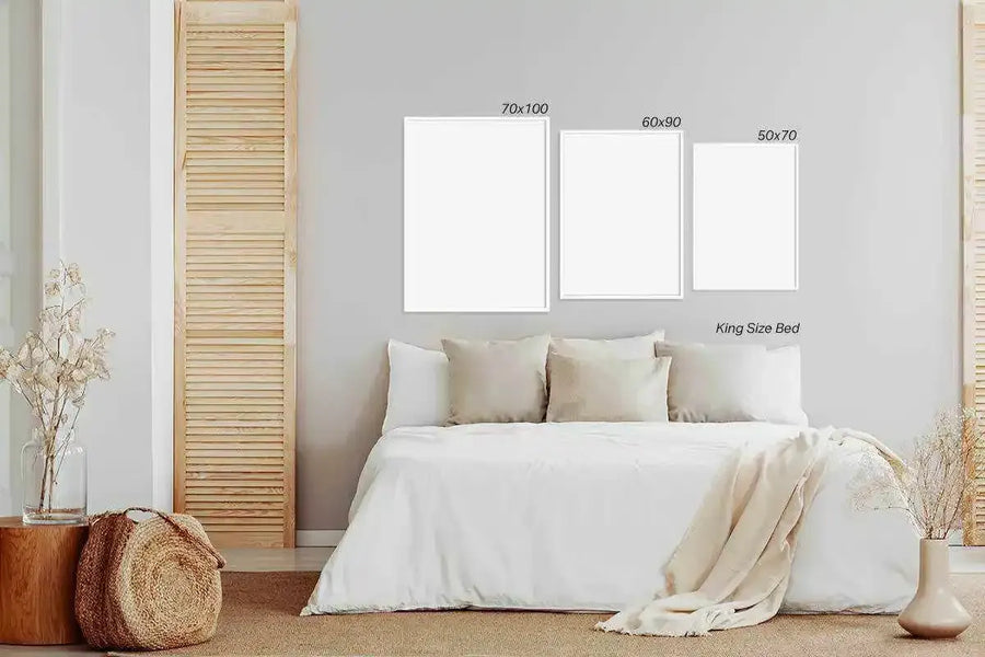 Lifestyle shot of three blank art prints displayed above a king-size bed in a minimalist bedroom. The prints are shown in three different sizes: 70x100, 60x90, and 50x70, all with thin white frames. The bed is covered with white bedding and several light beige pillows. To the left, a wooden stool holds a glass vase with dried flowers, and a woven basket sits on the floor. To the right, a light beige vase with dried flowers is visible.
