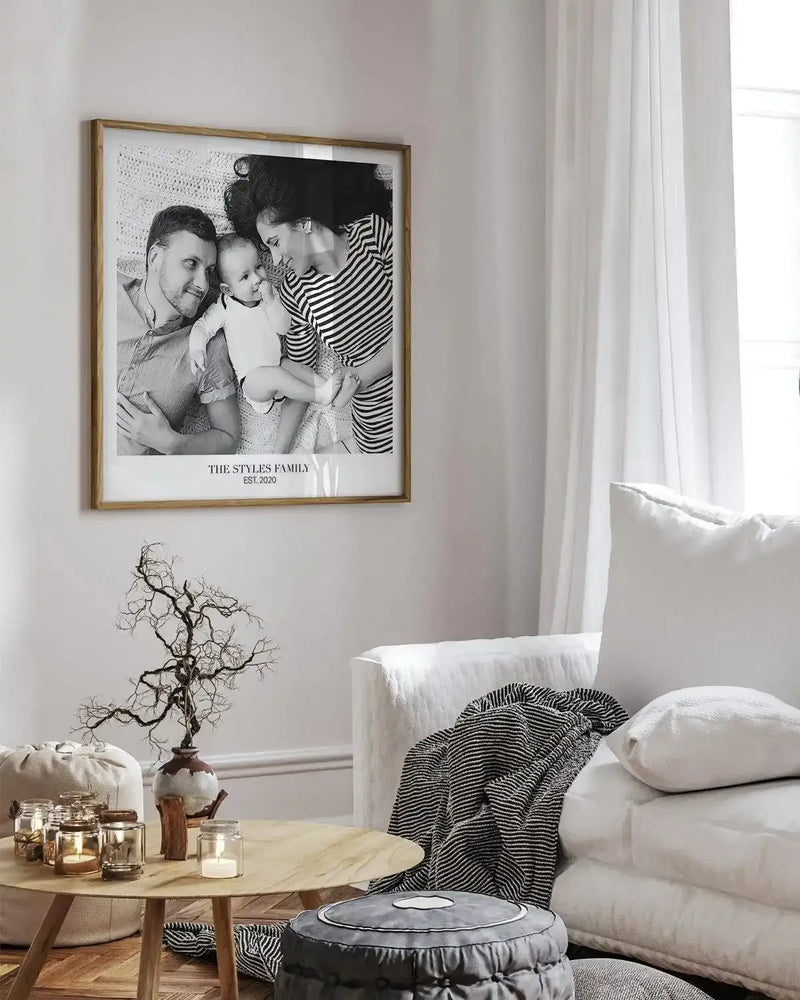 Large framed black and white photograph of a family displayed above a light wood coffee table in a minimalist living room. The print features a man, woman, and baby lying on a textured blanket, with the text "THE STYLES FAMILY EST. 2020" below. The room includes a white linen sofa with a striped throw, a dried branch arrangement in a ceramic vase, and various candles and poufs.