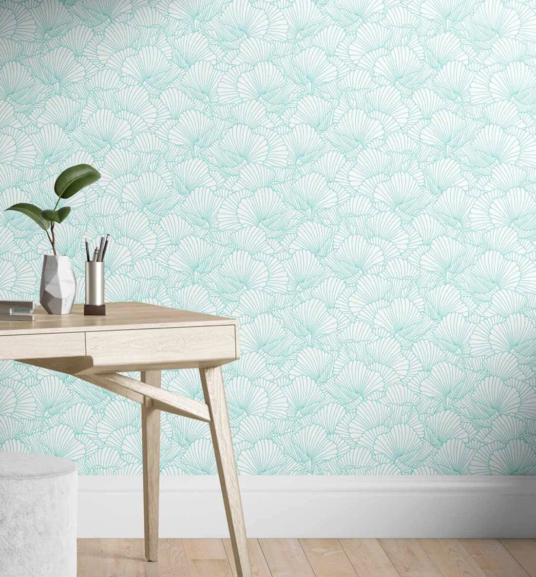 Lifestyle shot featuring Fan Coral Wallpaper in a home office setting. The wallpaper features a repeating pattern of delicate fan coral shapes in a soft, muted teal line art on a crisp white background. A light wood desk with a plant in a grey geometric vase and a silver pen holder sits against the wall.