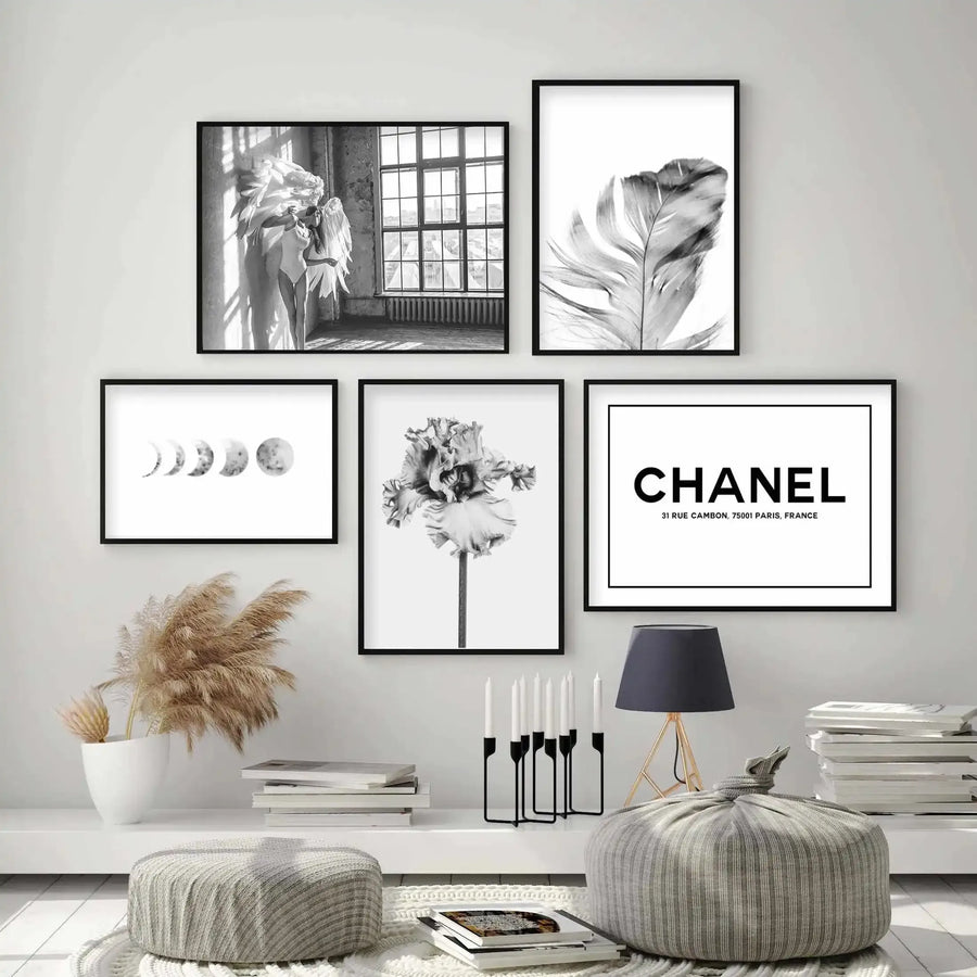 Black-framed grey art print featuring a delicate feather with detailed pencil-sketch shading against a white background. The print is displayed as part of a five-piece gallery wall arrangement in a minimalist room setting with neutral décor, including striped poufs, pampas grass, and stacked books, demonstrating how the feather artwork coordinates with modern home styling.