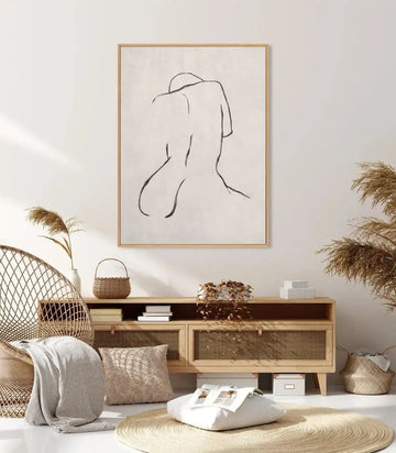 Lifestyle shot of a minimalist living room featuring a framed canvas art print of a female form in terra. The print depicts a simplified, continuous line drawing of a nude female figure from the back, seated with her head bowed, rendered in black on a cream background, displayed in a natural wood frame. The room includes a woven rattan chair with a striped cream throw, a light wood console table with woven doors, and dried pampas grass in a white vase and on the table.
