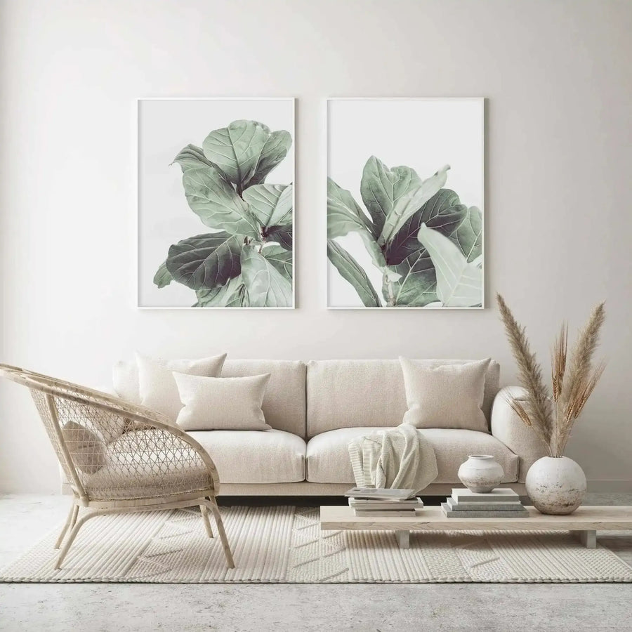 Two framed art prints displayed above a cream sofa in a minimalist living room. The prints feature close-up photographs of fiddle fig leaves in muted sage green and deep olive against a soft white background, with white frames and white borders. The room includes a woven rattan armchair, a light wood coffee table with books and ceramic vases, and dried pampas grass.