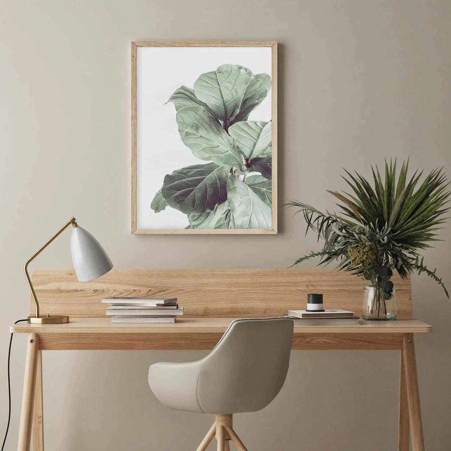 Lifestyle shot of a minimalist home office with a framed art print displayed above a light wood desk. The print features a close-up of large, textured fiddle leaf fig leaves in shades of muted sage green and deep emerald against a bright white background, with a natural wood frame and white border. The desk holds a gold and white desk lamp, a stack of books, and a glass vase with various green foliage. A light grey office chair is partially visible in the foreground.