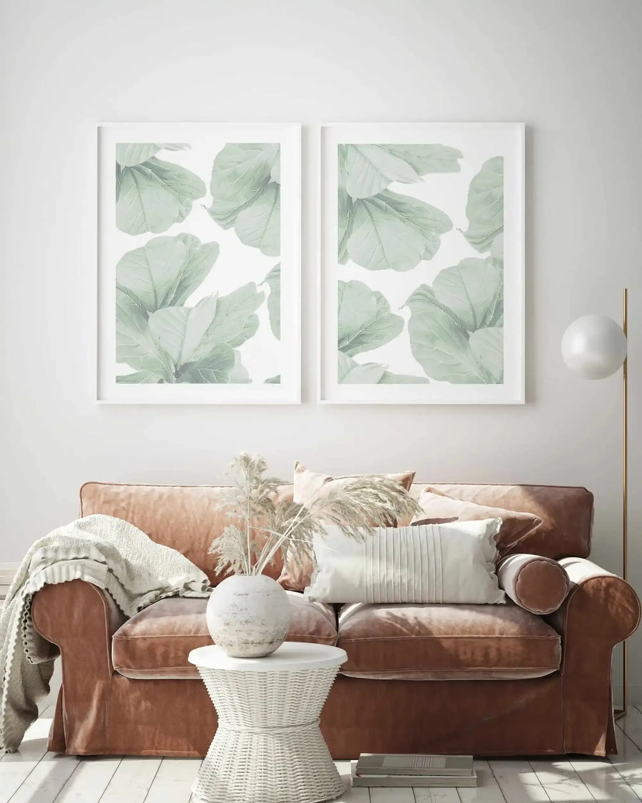 Two framed art prints displayed above a cognac-brown leather sofa. Each print features watercolor-style green fig leaves scattered across a white background, with soft sage and muted green tones showing delicate vein details. Prints are in white frames with white borders. Below, a white woven side table holds a decorative white ceramic vase with dried pampas grass, a cream throw blanket draped on the sofa arm, and neutral cushions create a calm, botanical-inspired living room setting.