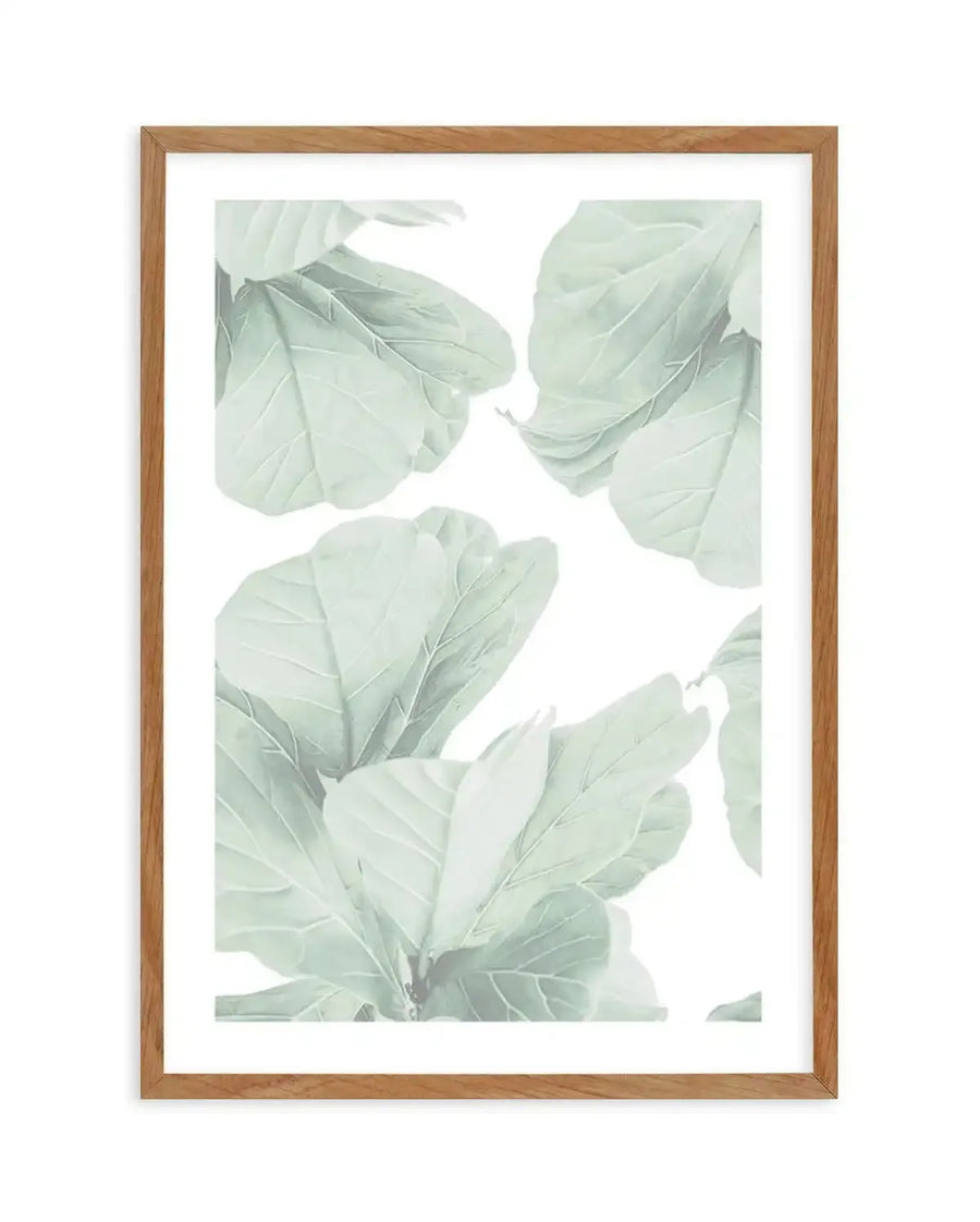 Framed art print featuring a watercolor illustration of pale sage green fig leaves arranged vertically against a white background. The leaves are rendered in soft, muted green tones with delicate vein details, creating a subtle, botanical aesthetic. The artwork is displayed in a natural wood frame with a white border. The composition shows overlapping leaves in varying sizes and positions, evoking a minimalist, nature-inspired design.