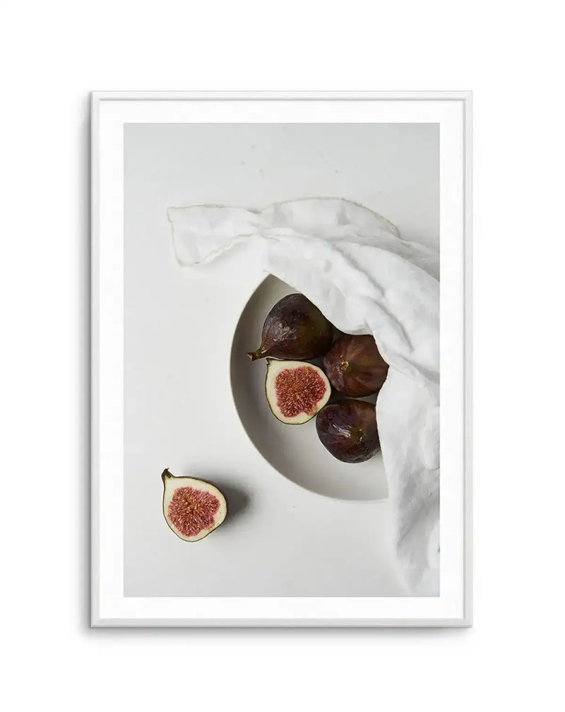 Framed photography art print of fresh figs on a white surface, displayed in a white frame with white border. The composition features a white bowl partially covered by a white linen cloth, holding several whole and halved dark purple figs with vibrant red-pink flesh and tiny seeds. One halved fig lies outside the bowl on the white surface. The image has a clean, minimalist aesthetic with soft shadows.